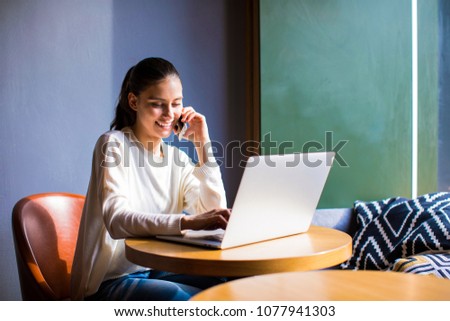 Cheerful woman associate web editor talking on mobile phone with customer and enter information in laptop computer, sitting in coffee shop. Smiling business female having cell telephone conversation 