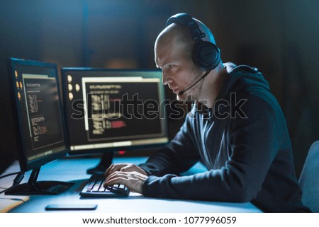 cybercrime, hacking and technology concept - male hacker with headset and coding on computer screen wiretapping or using computer virus program for cyber attack in dark room Royalty-Free Stock Photo #1077996059
