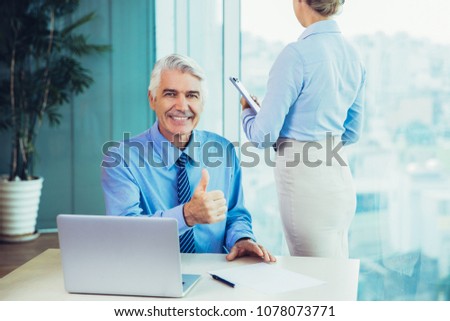 Smiling senior business man sitting at desk in office, showing thumb up and young female secretary standing near him back to camera with window and blurred city view outside in background