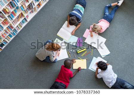 Elementary children lying on the floor and drawing at library. Top view of five multiethnic boys and girls in daycare house drawing on copybook. High angle view of group of kids with colorful pencils.