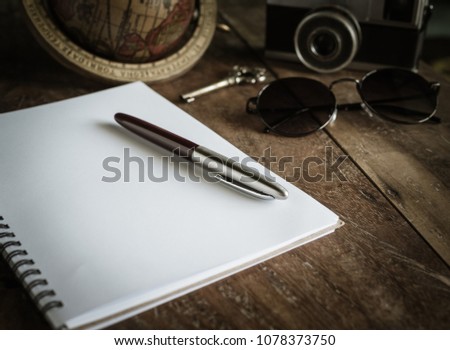 The Classic Silver Pen on The Blank White Notebook Paper with Antique Key, Sunglasses, Vintage Camera and The Classic World Globe on Old Wooden Backgroud for Traveller Journey Concept