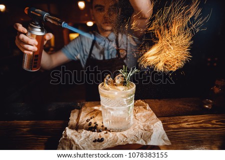 Barman prepares cocktail with orange and herbs in transparent glass on bar with alcohol. Uses burner with sparks. Dark background. Royalty-Free Stock Photo #1078383155