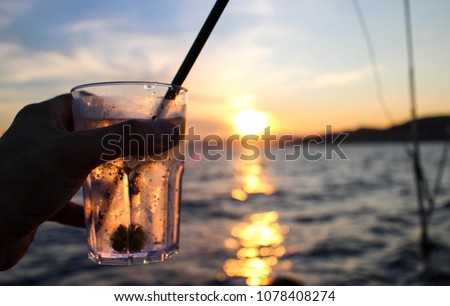 Glass of gin and tonic raised against the sunset over the sea. Sunlight reflected in the glass and costrasting the sea.