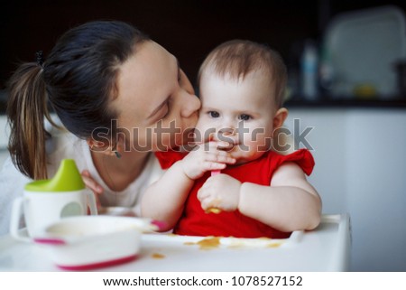 A cute baby in a red dress with plump cheeks sits on a white children's chair, eats a spoon from a plate, drinks from a cup. Young beautiful mother looks, smiles and kisses. Close-up.