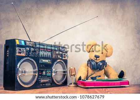 Retro Teddy Bear toy with headphones at the DJ turntable mixing console, classic mic, old cassette ghetto blaster radio recorder from 80s front concrete wall background. Vintage style filtered photo