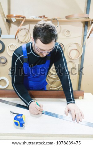 A bearded tired handsome carpenter on a table with a pencil draws a sign on the board. At the workplace there is an ordinary master dressed in blue cambenizon with a cruel haircut and saved glasses