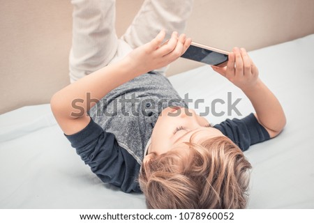 Small kid having fun and taking selfie with cell phone while lying on the bed. 