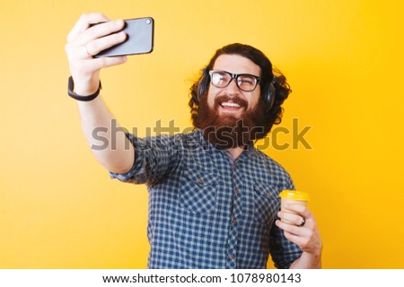 Cheerful bearded man taking a selfie and holding cup of coffee over yellow background