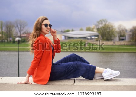 Beautiful girl wearing sunglasses at riverside. Girl sitting on pier and lookingat the river. Beautiful young girls outdoors in Sunny weather. Fashion model travel. Girl with long hair in red jacket.