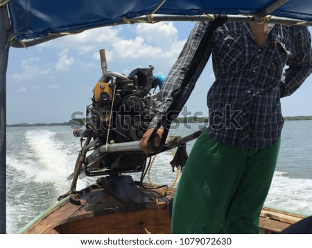 An unidentified man with dark skin wearing a checkered short and green pants drives a wooden motorboat with a blue synthetic roof in an ocean on a sunny day with background of cloudy blue sky.