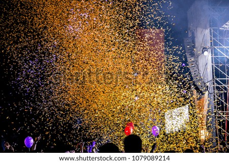 Hundreds of confetti fired on air during a festival at night. Image ideal for backgrounds. Orange tonality. People with the hands to the sky. Balloons and stage on the right
