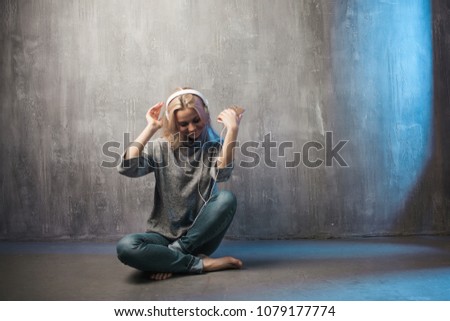 Young and attractive woman with a smartphone and large headphones, sitting on the floor. listening to music in the mobile app.Grey clothing and background