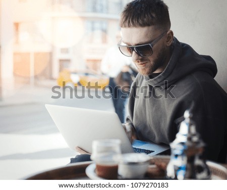 Pensive young man holding laptop on his knees and working on it in cafe. PC and man are on focus and background. Cutlery and tea are on foreground and blurred.