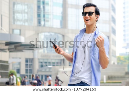 Hipster young man listening to music from mobile phone in city background. Happy caucasian man with cellphone, tourist on street, tourism, or summer holiday vacation travel concept.