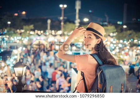 Portrait asian woman tourist backpacker and hat stand watch the night market with so many people for shopping fashion product in holiday and relax