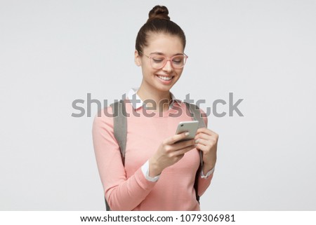 Indoor closeup of young European Caucasian girl pictured isolated on gray background with backpack on as if just returned from studies, looking at phone screen with nice smile, typing or browsing