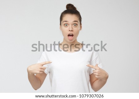 Indoor picture of young beautiful European woman isolated on grey background wearing white casual blank T-shirt that she is pointing to, showing deep astonishment with unexpected offer, copyspace