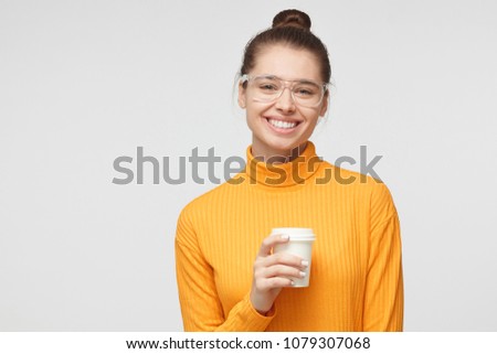 Closeup portrait of young beautiful woman pictured isolated on grey background in yellow top, having big eyeglasses on, holding cardboard cup of takeaway coffee, smiling happily, looking at camera