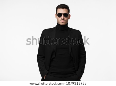 Half-length portrait of young handsome European guy isolated on white background dressed in total black with turtleneck, sunglasses and jacket, looking serious and suspicious, reserved and reticent Royalty-Free Stock Photo #1079313935