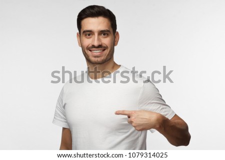 Horizontal picture of young handsome Caucasian man pictured isolated on grey background smiling positively while pointing with index finger to white blank T-shirt, copyspace for advertising