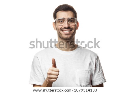 Closeup picture of young handsome European man pictured isolated on white background wearing transparent glasses and white T-shirt, showing thumb up as if sharing personal positive experience