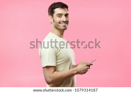Side portrait of young handsome European man isolated on pink background dressed in casual T-shirt, holding cellphone in hands and looking at camera while waiting for answer in social network