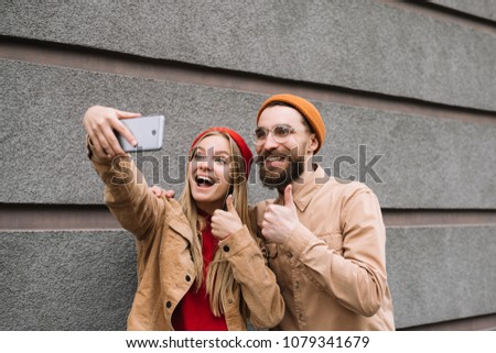 Positive and smiling hipsters friends taking selfies together, thump up. Stylish bearded man and beautiful blonde woman having fun, making pictures using smartphone on the street