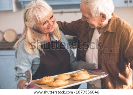 Portrait of happy mature woman taking care of her hungry husband. She is holding tray with cookies while man is embracing her with love