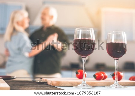 Focus on two glasses of red wine on table. Happy old loving couple dancing in kitchen on background