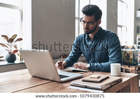                                Developing new project. Thoughtful young man writing something down while working in the creative working space Royalty-Free Stock Photo #1079430875