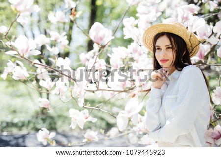 Beauty woman in straw hat standing on Magnolia blossoming flowers background in spring sunny day