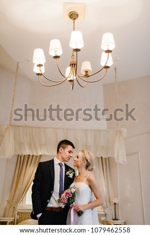 Young bride and groom standing and kissing indoors in studio or home with a large chandelier. Portrait woman and man posing in interior.