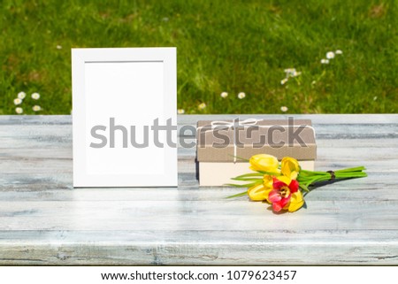 Gift box, flowers and white vintage frame with place for your text on rustic bright wooden table. Green natural background. Card concept. Outdoor.