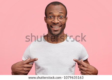 Portrait of handsome cheerful dark skinned male seller of clothes, dressed in casual white t shirt, indicates at blank copy space for your logo, isolated on pink background. People, clothing, design