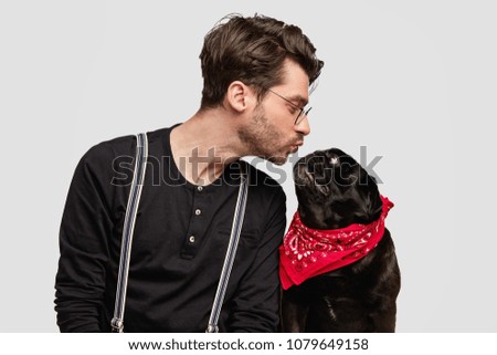 Sideways shot of affectionate dog`s male host going to kiss his lovely black pug dog with red bandana, expresses his love and devotion. People, animals, loyalty and truthful friendship concept