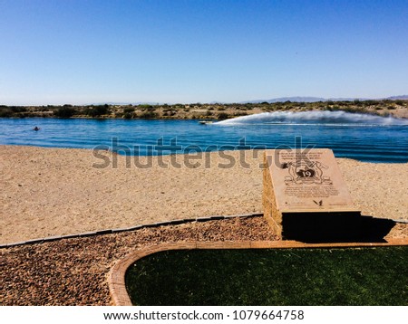 A memorial plaque on the shoreline for the Granite Mountain Hotshots next to a sandy beach, as the Colorado River floats by in the background and a boat speeds by shooting out a large roostertail