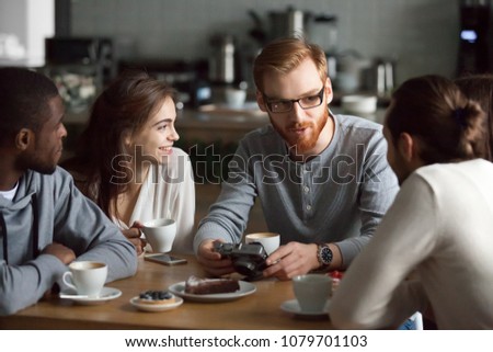 Young redhead guy holding camera showing photos sharing recent memories or travel impression hanging with diverse friends, smiling multiracial millennial people talking sitting together at cafe table