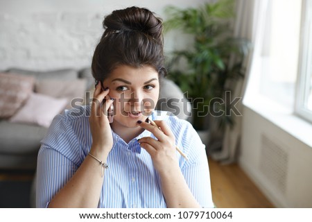 Stylish chubby student girl with messy hair bun talking to friend on mobile while doing homework. Picture of attractive young brunette overweight female counsellor making appointment using cell phone