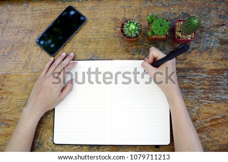 Top view, A female hands holding a pen writing notebook on the wooden table with smartphone and three cactus. 