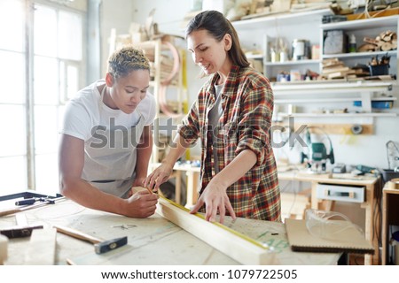 Two young intercultural women measuring length of wooden plank before sawing it on workbench Royalty-Free Stock Photo #1079722505