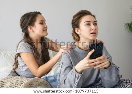 Horizontal shot of cute girl with long wavy hair sitting in bed, disturbing busy mother with mobile, shaking her by shoulders, feeling impatient while wanting to play online video games on cell phone