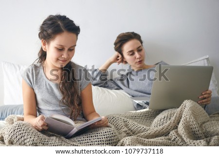 Ideal cozy picture of charming 10 year old female child enjoying favorite fairytale, holding book while sitting in bed with her mother who is working remotely using portable computer in background