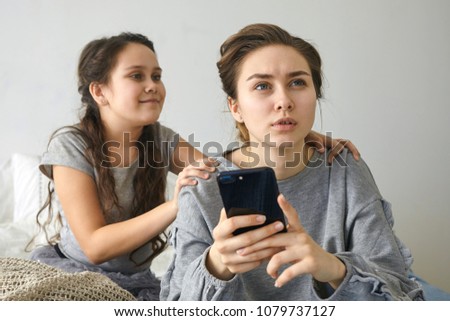 Picture of attractive serious young woman in casual wear holding mobile phone, having concentrated look, making calculations while doing finances at home, her naughty daughter disturbing her