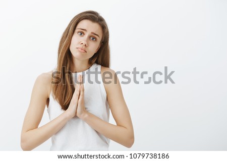 Please, forgive me darling. Portrait of timid cute caucasian female coworker in casual outfit, holding hands in pray, lifting eyebrows and tilting head while asking apology or favor, begging for help