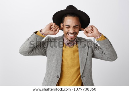 Using natural earplugs not to hear spoilers. Good-looking happy dark-skinned guy in classy hat and stylish outfit smiling with closed eyes, holding index fingers in ears, waiting for surprise Royalty-Free Stock Photo #1079738360