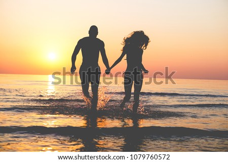 Couple of lovers walking inside water on tropical beach in summer vacation at sunset - Young people enjoying holidays - Love, travel and landscape concept - Focus on silhouettes Royalty-Free Stock Photo #1079760572