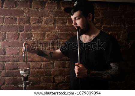 Young hookah maker adjusts coals. Tatooed hookah server. Brick wall in the background.