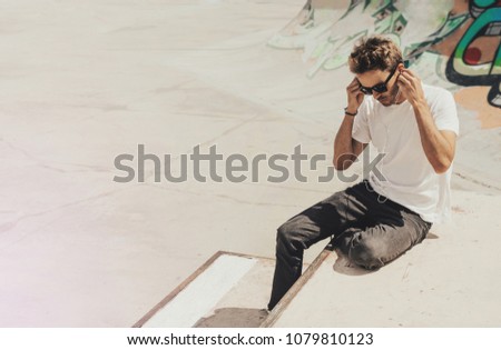 Young handsome bearded man sitting next to a graffiti wall background in skate park is wearing a white blank t-shirt and sunglasses. Horizontal mock up style.