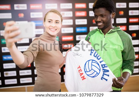 Portrait of smiling African-American sportsman taking selfie with fan and holding signed t-shirt