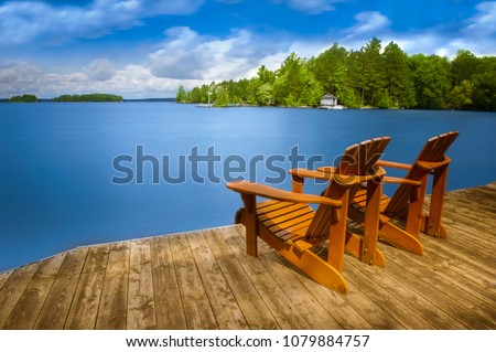 Two Adirondack chairs sitting on a wooden dock facing a blue calm lake. Across the water is a white cottage nestled among green trees.  Royalty-Free Stock Photo #1079884757